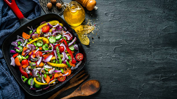 Vegan food: grilled vegetables in a cooking pan. Overhead view with copy space. Vegan food backgrounds: overhead view of a cast iron cooking pan filled with grilled vegetables shot on dark kitchen table. The composition is at the left of an horizontal frame leaving useful copy space for text and/or logo at the right.
Vegetables and spices included in the composition are bell pepper, tomatoes, celery, broccoli, onion, carrots, corn, rosemary, dried parsley, peppercorns, salt, among others. An olive oil bottle complete the composition. High resolution 42Mp studio digital capture taken with Sony A7rII and Sony FE 90mm f2.8 macro G OSS lens chopped food photos stock pictures, royalty-free photos & images