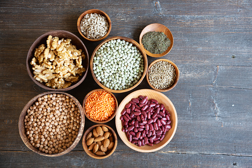 A variety of plant based proteins like legumes, nuts and seeds in wooden bowls on a dark wooden background, seen from above. These vegan proteins are: almonds, walnuts, sunflower seeds, hemp seeds, nettle seeds, red lentils, kidney beans, chick-peas, dried green peas