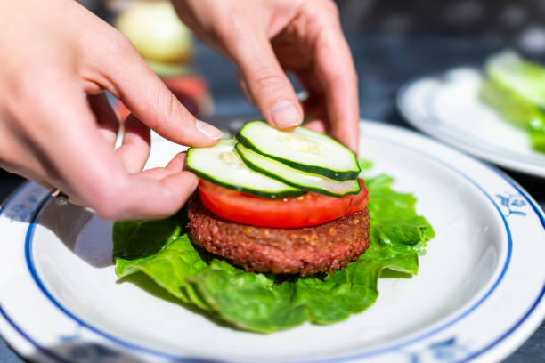 vegan vlees worst patty op plaat met romaine sla blad en tomaten komkommers gesneden voor hamburger met vrouwelijke hand regelen van schotel - vegan keto stockfoto's en -beelden
