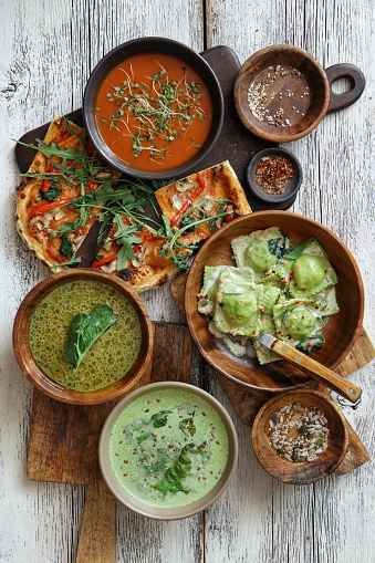 Vegan spinach ravioli, nuts, seeds and herb sauce. Vegetarian pizza with tomato sauce, spinach, mushroom, arugula, bell pepper, sun-dried tomatoes, seeds and pesto. Roasted red bell pepper soup. Minestrone with pesto. Spinach soup. Flat lay top-down composition on white wooden background.