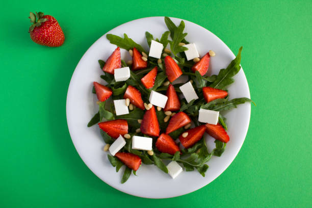 Vegatarian salad with strawberry, arugula, soft cheese  and pine nuts  in the white plate on the green background. Top view. stock photo