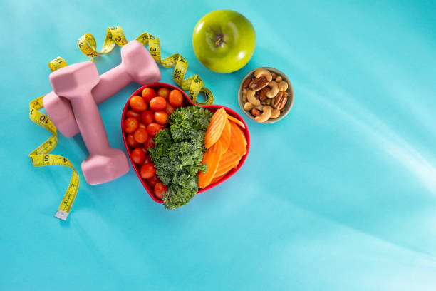 This is an overhead photograph of vegetables in a heart bowl  sitting next to dumbells, nuts and a green apple on a blue background. This image symbolizes healthy living and eating lifestyle. There is a lot of copy space on the right