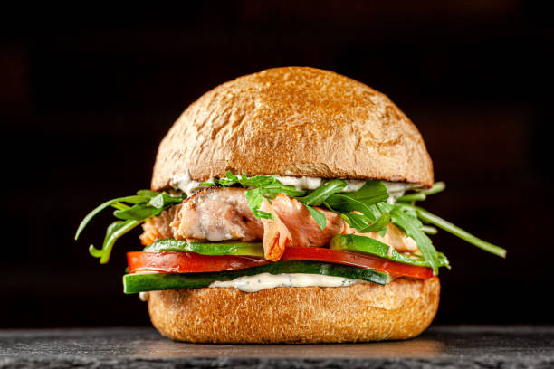 Vegetarian American Cuisine. Burger with red fish salmon, arugula, cucumbers and tomatoes, white sauce. Burger on a black background. background image, copy space text stock photo
