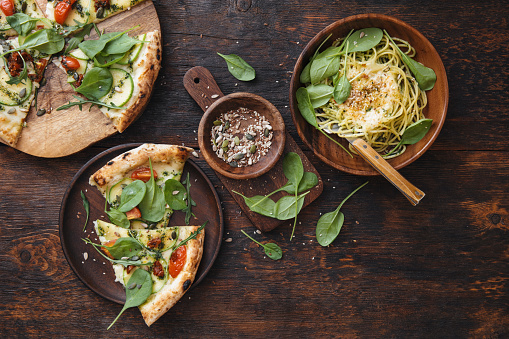 Vegetarian spaghetti with zucchini, spinach, seeds, vegan cheese and pesto sauce. Vegetarian pizza with zucchini, sun-dried tomatoes pesto sauce , spinach and nuts. Flat lay top-down composition on dark wooden background.