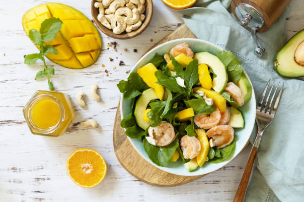 Vegetarian vegan healthy food. Salad with arugula, mango, avocado, shrimp, pecans and dressing of olive oil, honey and wine vinegar on a white rustic kitchen table. Top view, flat lay background. stock photo