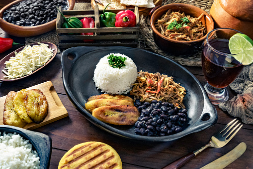 Venezuelan traditional food, Pabellon Criollo with arepas, casabe and papelon with lemon drink. Home made with white rice, black beans, fried plantains, and shredded beef. Plate on a wooden table in a rustic kitchen. Set with ingredients and shredded white cheese. Pabellon Criollo, is a main meal in the Venezuelan Culture and cuisine.