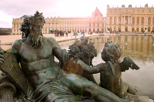 Paris, France - January 11, 2010: Gardens of Versailles. Statue on the Water Parterre at sunset. The palace facade and tourists enjoying can be seen in the background. This is one of the most visited monuments in France.