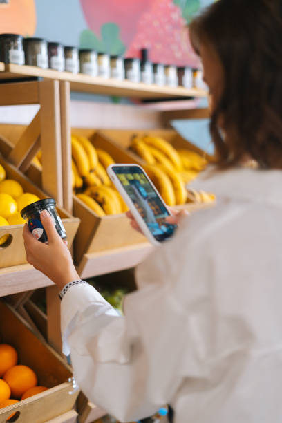 Vertical cropped shot of unrecognizable young woman choosing products in market walking along counters taking photos of items using smartphone. stock photo