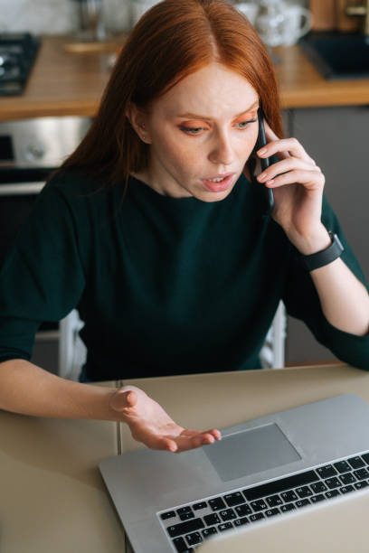 Vertical shot of arguing angry young woman talking on mobile phone and using laptop sitting at table in kitchen with modern interior. stock photo