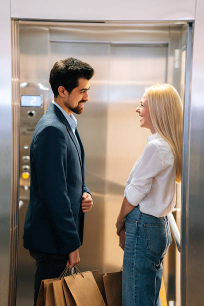 Vertical shot of cheerful young man and beautiful woman looking at each other smiling standing inside of elevator in shopping mall. stock photo