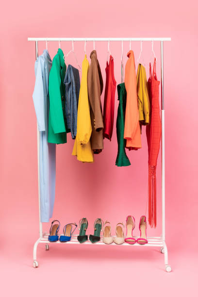 Vertical Shot Of Clothing Rail With Clothes Over Pink Background Shopping Concept. Vertical Shot Of Clothing Rail With Bright Trendy Clothes Hanging On Hangers And Shoes Over Pink Studio Background In Empty Room. Fashion Trends And Style Concept clothes rack stock pictures, royalty-free photos & images