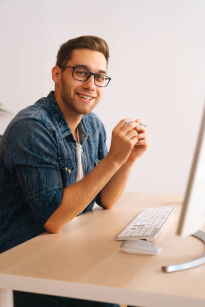 Vertical side view shot of smiling handsome young freelance designer male in stylish glasses working on desktop computer holding pen in hands stock photo