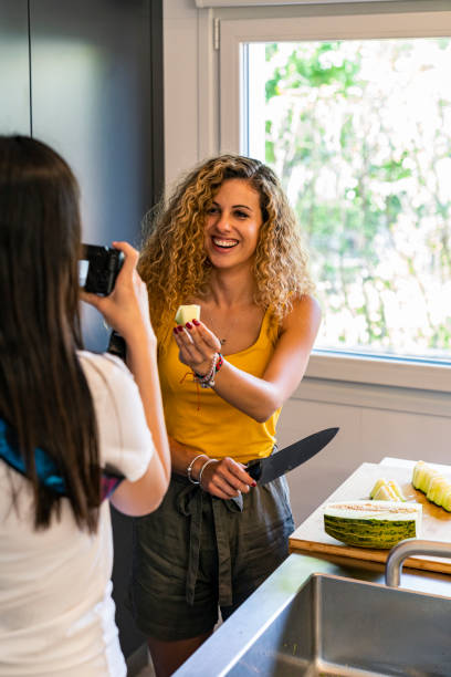 vertical stock photo of a two girls of different ethnicities in the kitchen, one shows a piece of melon to the camera of the other, which shoots a photo. holidays and friends - asiatic kids room imagens e fotografias de stock