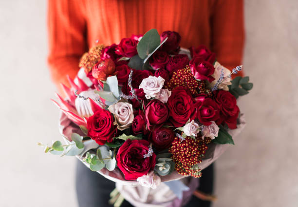 très belle jeune femme en chandail rouge tenant fleur bouquet de fleurs de roses fraîches, oeillets, eucalyptus dans des couleurs vives de passionnés rouges sur le fond du mur gris - bouquet de fleurs photos et images de collection