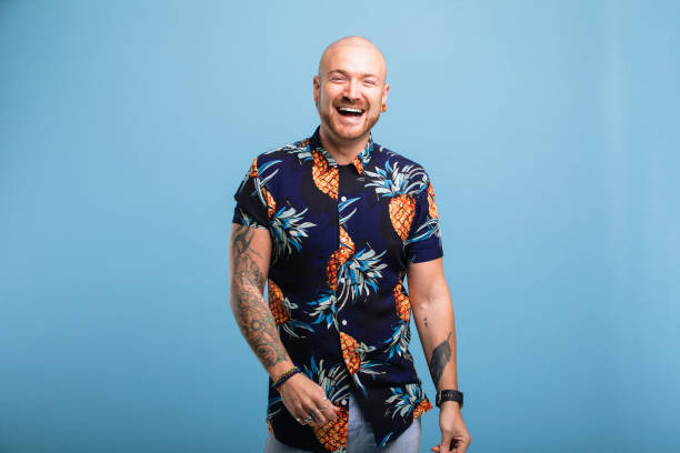 パイナップルプリントのシャツを着て、笑顔でカメラを見て入れ墨を持つ中年の男性の肖像画。彼は青いスタジオの背景の前に立っています。
