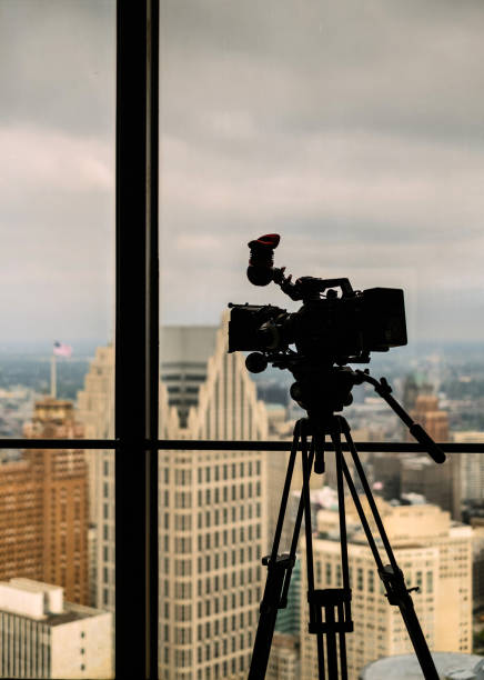 Video camera ready for shoot Conture of modern professional 4K video camera on tripod against the window view of modern cityspace with office buildings and moody sky in the background. michigan shooting stock pictures, royalty-free photos & images