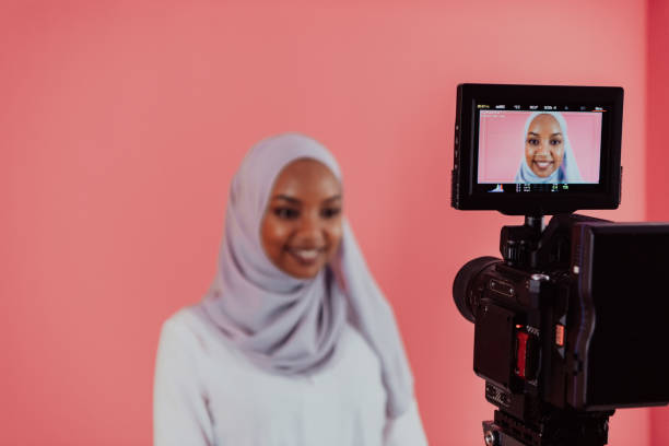 Videographer in digital studio recording video on professional camera by shooting female Muslim woman wearing hijab scarf plastic pink background stock photo