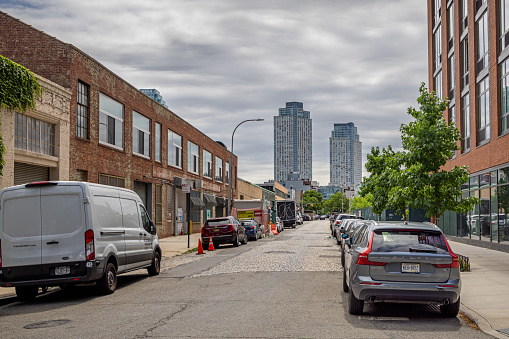 Long Island City, Queens, New York, NY, USA - July 7th 2022: View down 5th Street with parked cars, small businesses and cobblestones toward two modern skyscrapers