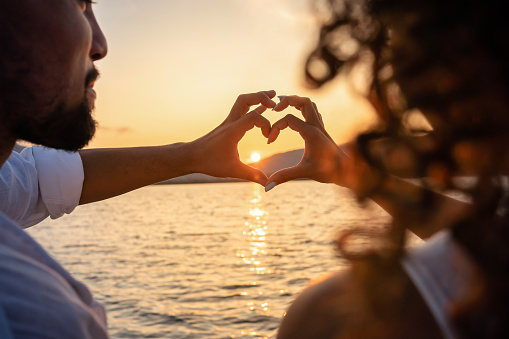 View from back of a young beautiful couple doing heart shape with fingers looking at the sun setting behind the mountains, reflecting light in the sea water. Valentine day love concept. Focus on hands