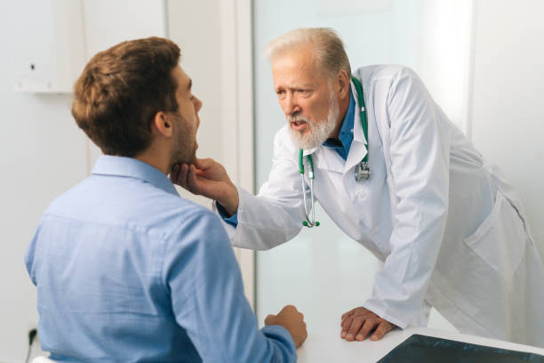 View from back to mature adult male doctor otolaryngologist performing examination of young man patient throat in office of medical clinic. stock photo