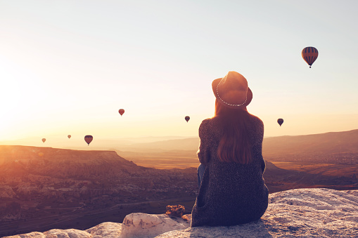 View from the back of a girl in a hat sits on a hill and looks at air balloons in Cappadocia in Turkey.