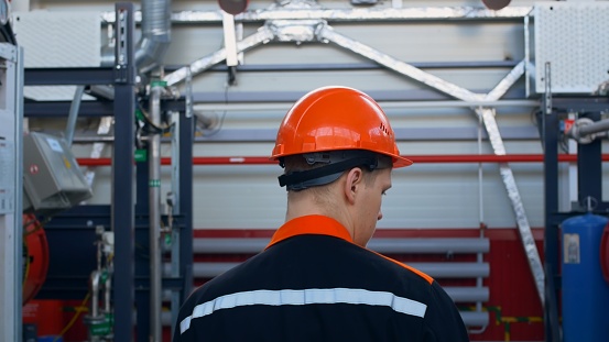 A view from the back of a worker in an orange helmet and overalls walks around his equipment at a chemical plant in the pump shop, checking the condition of the equipment. Factory work.