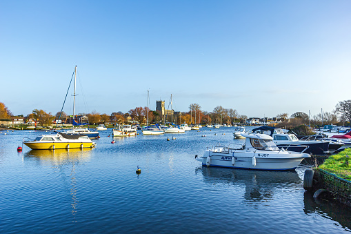 A view of a beautiful calm river marina with boats along grassy banks and church in the background under a majestic blue sky