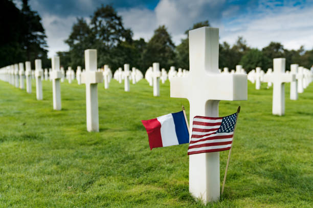 вид на крест надгробия на американском кладбище на пляже омаха в нормандии с французскими и американскими флагами - colleville стоковые фото и изображения