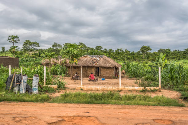 View of traditional village, two women and a child sitting in front of thatched house with roof and terracotta and straw walls, cloudy sky as background stock photo