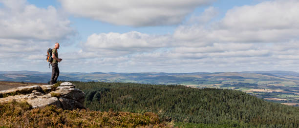 A View Worth the Hike A senior man wearing a backpack on a hike in the hills in Rothbury, Northumberland. He is standing on a cliff edge and looking at the beautiful green scenery. rothbury northumberland stock pictures, royalty-free photos & images