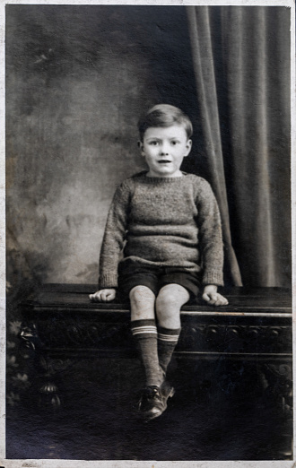 Vinatge photo of a small boy, aged 5 years. Circa 1933. Interior shot with the child seated on an ornate wooden bench, with draped curtain background.