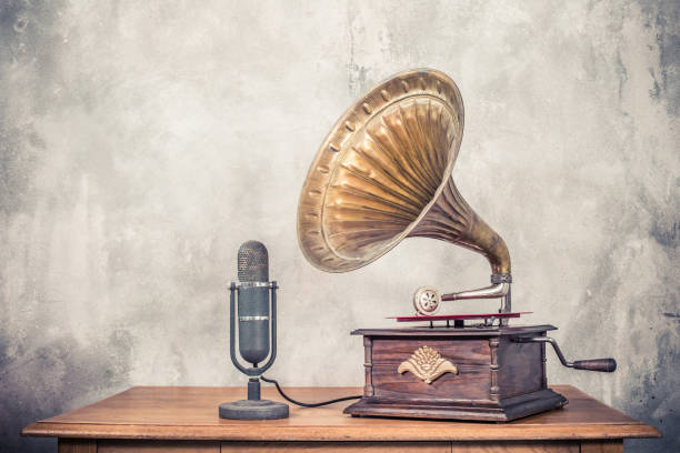 Vintage antique gramophone phonograph turntable with brass horn and big aged studio microphone on wooden table front concrete wall background. Retro old style filtered photo stock photo