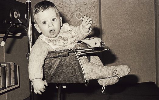 Vintage black and white image of a cute and surprised baby boy on a baby chair looking straight to camera .