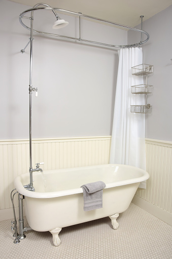A classic Victorian style cast iron claw foot bathtub with water running from tap in an old fashioned bathroom with beadboard walls and tiled floor.