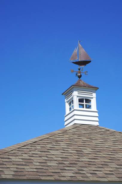 Vintage sailing ship weather vane atop a white cupola on top of shingled gable roof with blue sky background Iconic metal ship weather vane mounted on classic New England rooftop with square white cupola and brilliant blue sunny sky cupola stock pictures, royalty-free photos & images