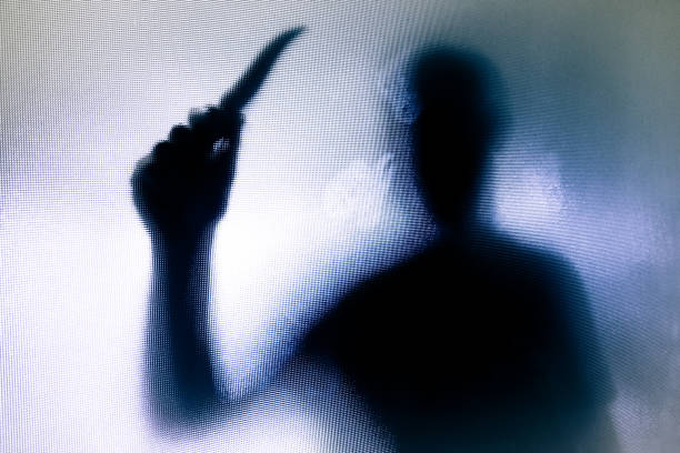 Violent threatening silhouette of man wielding a knife behind frosted glass window Monochrome backlit image of the silhouette of a man wielding a sharp knife in an aggressive way. The silhouette is distorted, and the arms elongated, giving an alien-like quality. The image is sinister and foreboding, with an element of horror. The image conveys a domestic violence, knife crime theme. Horizontal image with copy space. murder stock pictures, royalty-free photos & images