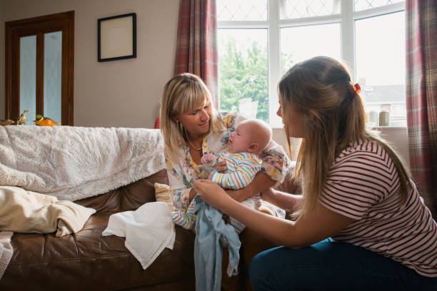 Visiting Her Newborn Grandson Mid adult mother is at home with her baby son. His grandmother is visiting and helping to look after the baby as he is crying. visit stock pictures, royalty-free photos & images