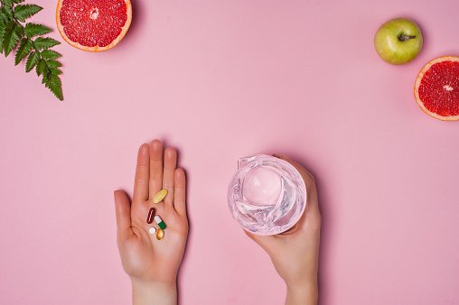 Vitamins and a glass of water in female hands on a pink background. Place for text. Grapefruit, apple and green leaf on the background. Healthy lifestyle concept