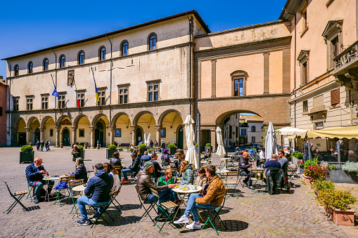 Viterbo, Italy, April 18 -- Customers and tourists enjoy life in Piazza del Plebiscito in the historic heart of the medieval city of Viterbo, in central Italy. In the background, the Palazzo dei Priori, built in 1264 and also used as the seat of the Pontifical Government. In 1460 the building took on its current Renaissance style structure and is currently the Town Hall of Viterbo. The medieval center of Viterbo, the largest in Europe with countless historic buildings, churches and villages, stands on the route of the ancient Via Francigena (French Route) which in medieval times connected the regions of France to Rome, up to the commercial ports of Puglia, in southern Italy, to reach the Holy Land through the Mediterranean. Founded in the Etruscan era and recognized as a city in the year 852, Viterbo is characterized by its peperino stone and tuff constructions, materials abundantly present in this region of central Italy. Super wide angle image in high definition format.