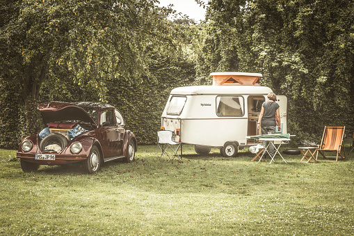 Volkswagen Beetle classic car  with a small caravan in a field with fruit trees. A woman is setting up the camping gear in front of the caravan. The car is on display during the 2016 Classic Days at Schloss Dyk in Juchen, Germany.