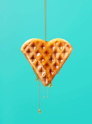 Pouring maple syrup over one heart-shaped waffle against a green-colored background. Close-up of maple syrup dripping on a waffle.
