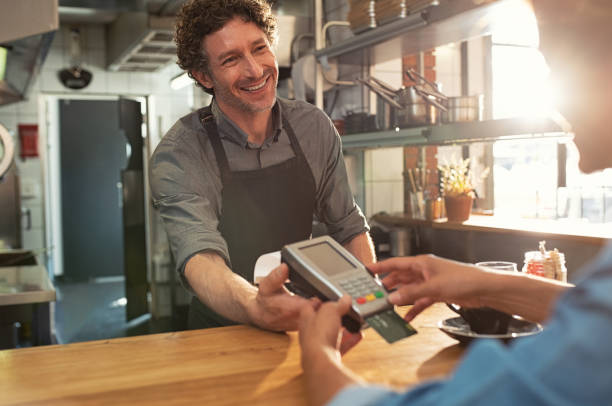 Waiter accepting payment by card Woman paying by credit card and entering pin code on reader holded by smiling barista in cafeteria. Customer using credit card for payment. Mature cashier wearing apron accepting payment over nfc technology. credit card reader stock pictures, royalty-free photos & images