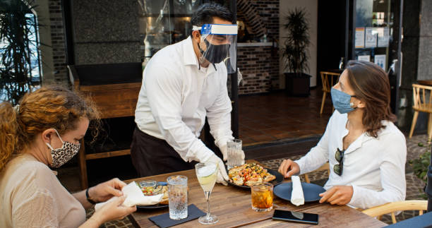 Waiter Wearing PPE During Covid-19 Pandemic Serving Food to Diners Wearing Masks A high end restaurant reopens after the 2010 Covid-19 lockdown, with measures in place to protect staff and customers. outdoor dining stock pictures, royalty-free photos & images