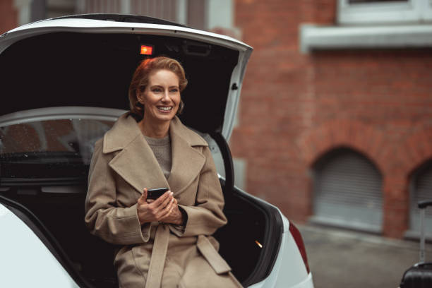 Waiting for her husband A 40-year-old German woman is waiting for her husband. She is sitting on the trunk of their car and holds the smartphone in hands. georgijevic frankfurt stock pictures, royalty-free photos & images