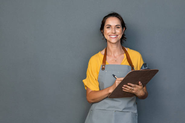 Waitress in apron take orders Happy smiling waitress taking orders isolated on grey wall. Mature woman wearing apron while writing on clipboard standing against gray background with copy space. Cheerful owner ready to take customer order while looking at camera. Small business concept. waiter taking order stock pictures, royalty-free photos & images