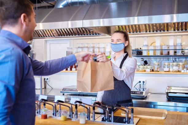 Waitress wearing protective face mask serving takeaway food to customer at counter in small family restaurant. Young waitress waring protective face mask and apron serving customer at counter in small restaurant - Small business and entrepreneur concept with woman owner in eatery with takeaway service delivery restaurant environment stock pictures, royalty-free photos & images