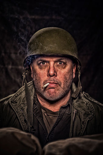 War Worried Vintage Army Soldier, in a fox hole, looking at the camera, smoking a cigarette,  behind sand bags, against a dark background