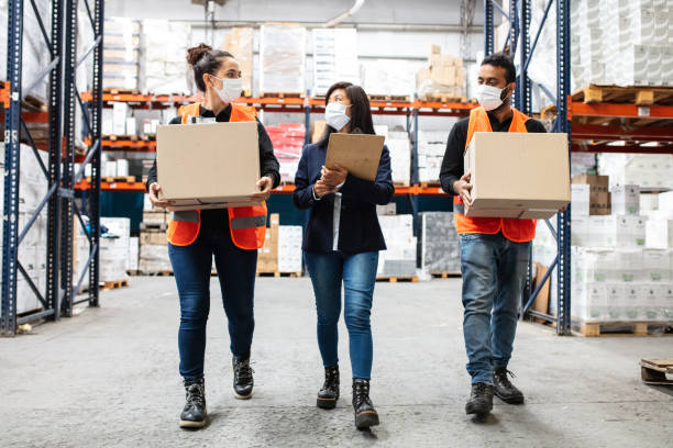 Warehouse manager with workers working during pandemic Warehouse manager walking with two employees carrying cardboard boxes. Businesswoman with male and female workers wearing face masks in warehouse. indian women walking stock pictures, royalty-free photos & images