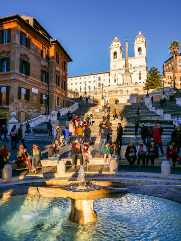 Rome, Italy, February 10 -- A warm ray of sun at sunset illuminates the Fontana della Barcaccia at the foot of the monumental stairway of Trinità dei Monti, or Spanish Steps, in the baroque heart of Rome, with dozens of tourists around. This iconic fountain in Rome was finished in 1629 by the architect and sculptor Pietro Bernini, father of the more famous Gian Lorenzo Bernini, commissioned by Pope Urban VIII. According to tradition, Gian Lorenzo himself participated in the final construction of the fountain on the death of his father in 1629. A masterpiece of the late Roman Baroque, the monumental stairway of the Trinità dei Monti (Spanish Steps) was built in 1721 to connect the church of the Santissima Trinità dei Monti (top in the photo) with the Piazza di Spagna (Spanish Square). Designed by the architect and urban planner Francesco De Sanctis, the Spanish Steps were inaugurated by Pope Benedict XIII on the occasion of the Jubilee of 1725. Built starting from 1502 in the Renaissance style, the church of the Santissima Trinità dei Monti was commissioned by Charles VIII, King of France, to give a stable accommodation to the religious Order of the Minims. In 1570 the façade was rebuilt based on a design by the architects Giacomo Della Porta and Carlo Maderno, with the construction of the two characteristic bell towers. In 1980 the historic center of Rome was declared a World Heritage Site by Unesco. Super wide angle image in high definition format.