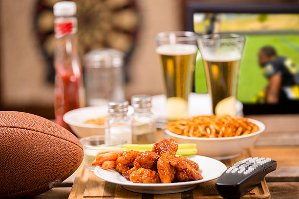 Hot wings and football in foreground.  Beer in mugs in background with television.  Football game on TV in a local pub or sports bar.  Dartboard in background.  Bar top. championship game party!